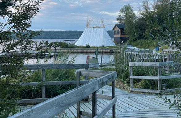 Passerelle en bois bordée de végétation menant à un site au bord de l’eau, avec un grand tipi et un bâtiment en arrière-plan, sous un ciel partiellement nuageux.